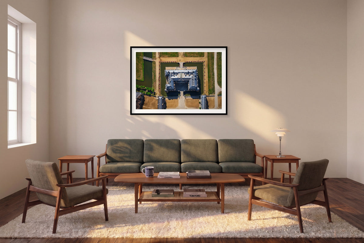 Overhead view of a French château surrounded by a green moat, pale paths, bridges and pink flowers in the Paris region; black framed print in a room setting