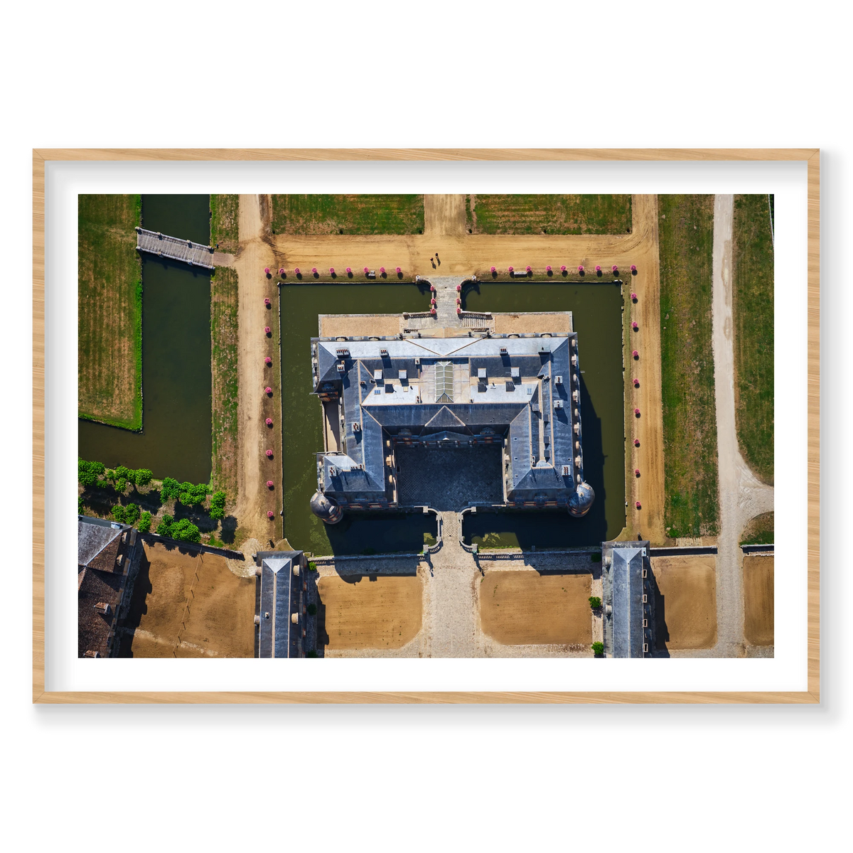 Overhead view of a French château surrounded by a green moat, pale paths, bridges and pink flowers in the Paris region; natural timber frame