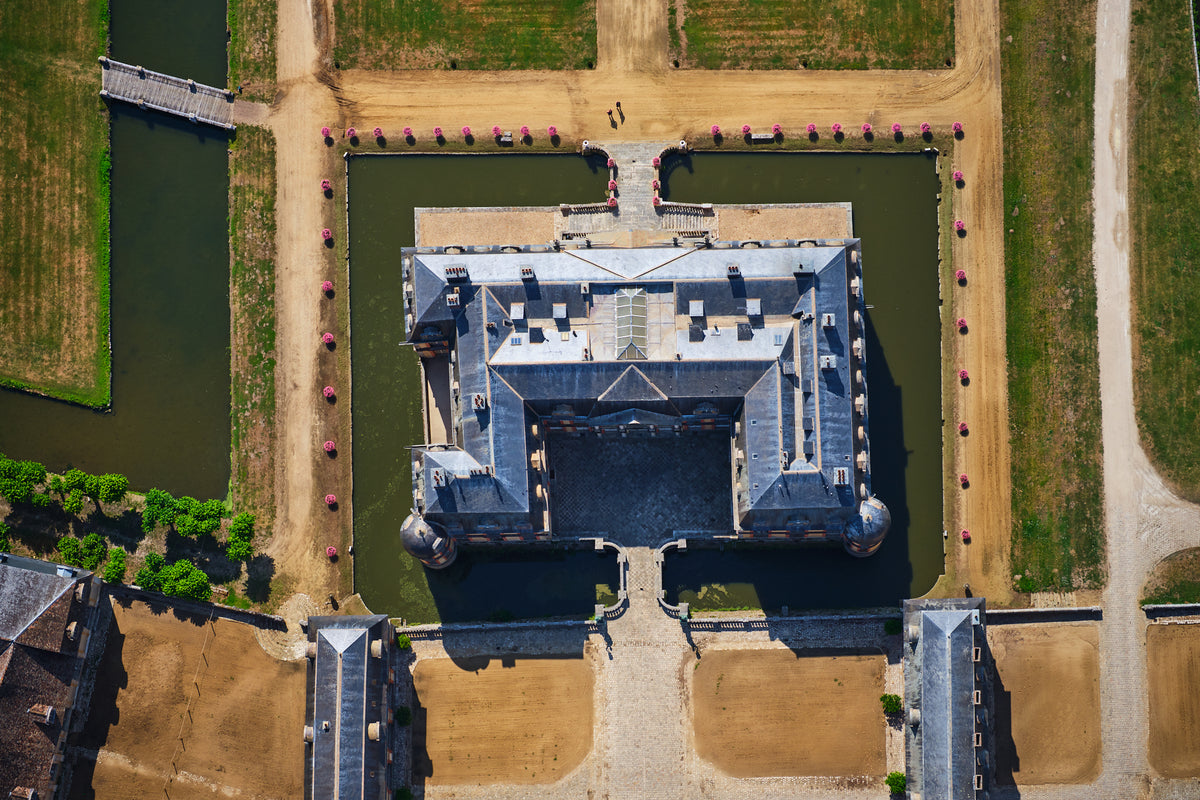 Overhead view of a French château surrounded by a green moat, pale paths, bridges and pink flowers in the Paris region; print only