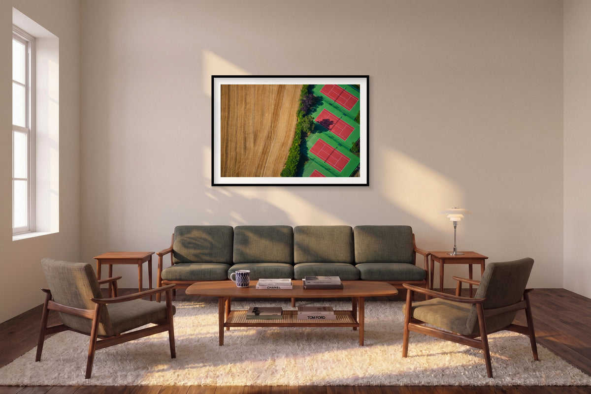 Red tennis courts with green surrounds beside a golden field, seen from above in the Paris region, France; black framed print in a room setting