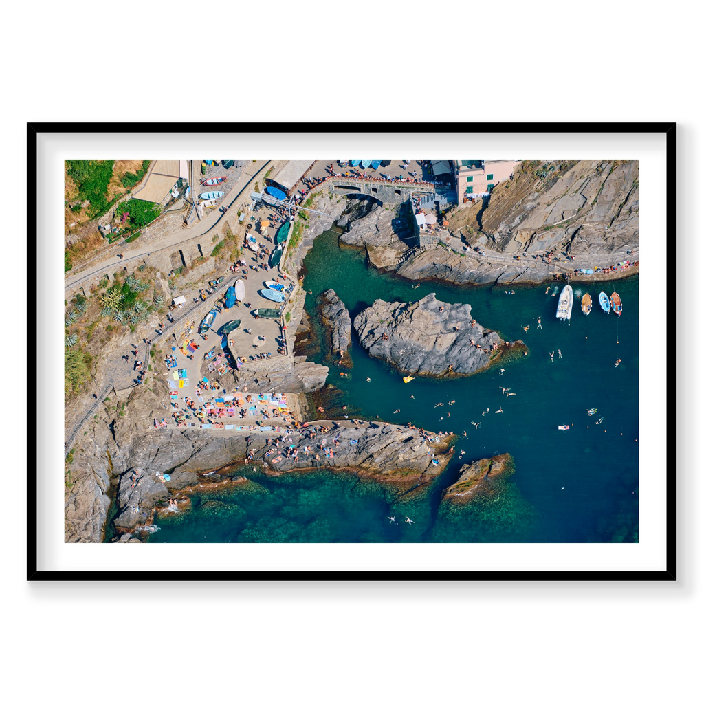 Overhead view of swimmers, boats and sunbathers around Manarola’s rocky harbour, Cinque Terre, Italy, black frame.