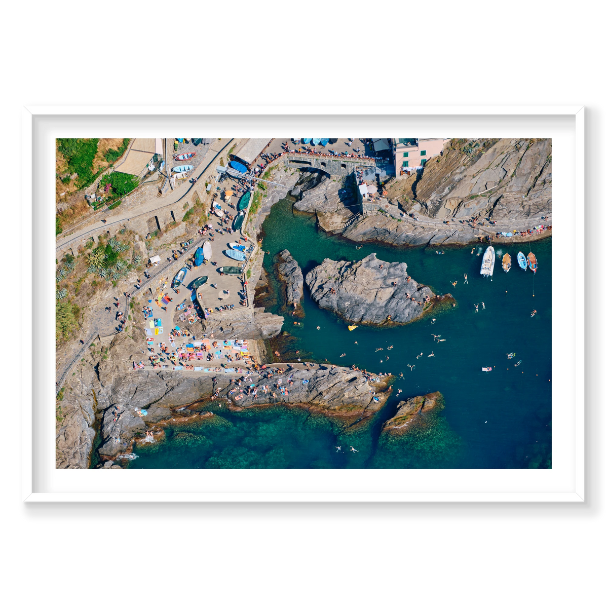Overhead view of swimmers, boats and sunbathers around Manarola’s rocky harbour, Cinque Terre, Italy, white frame.