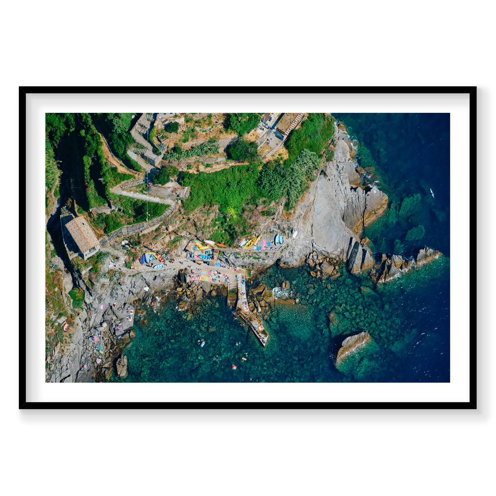 Overhead view of winding hillside steps, colourful boats, a rocky landing and swimmers in clear water at Corniglia, Cinque Terre, Italy, black frame.