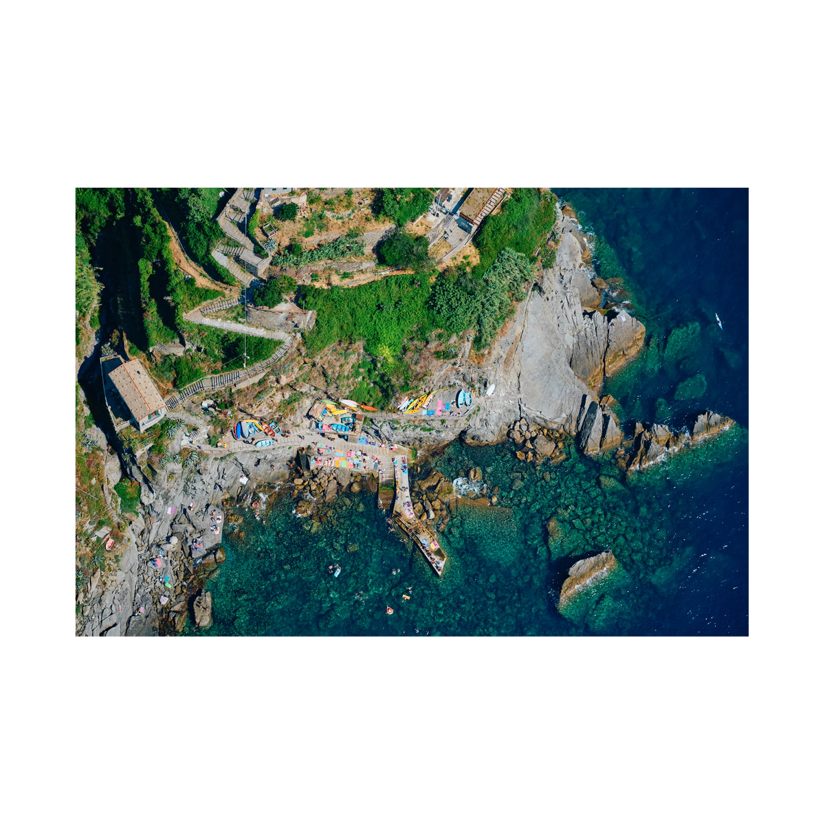 Overhead view of winding hillside steps, colourful boats, a rocky landing and swimmers in clear water at Corniglia, Cinque Terre, Italy, print-only presentation.