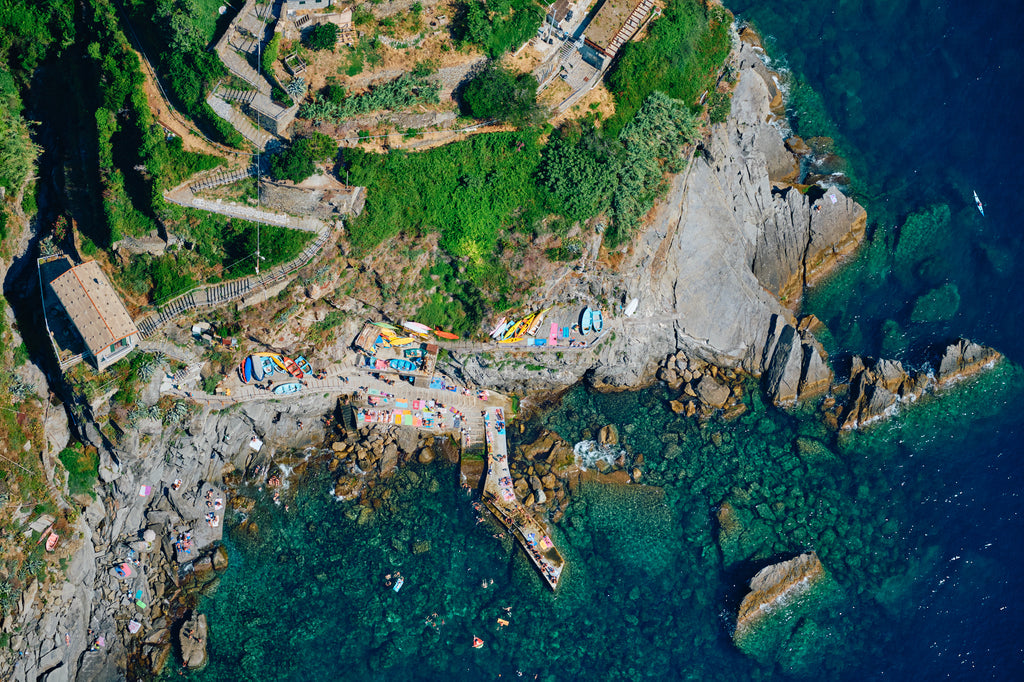 Overhead view of winding hillside steps, colourful boats, a rocky landing and swimmers in clear water at Corniglia, Cinque Terre, Italy, print-only presentation.