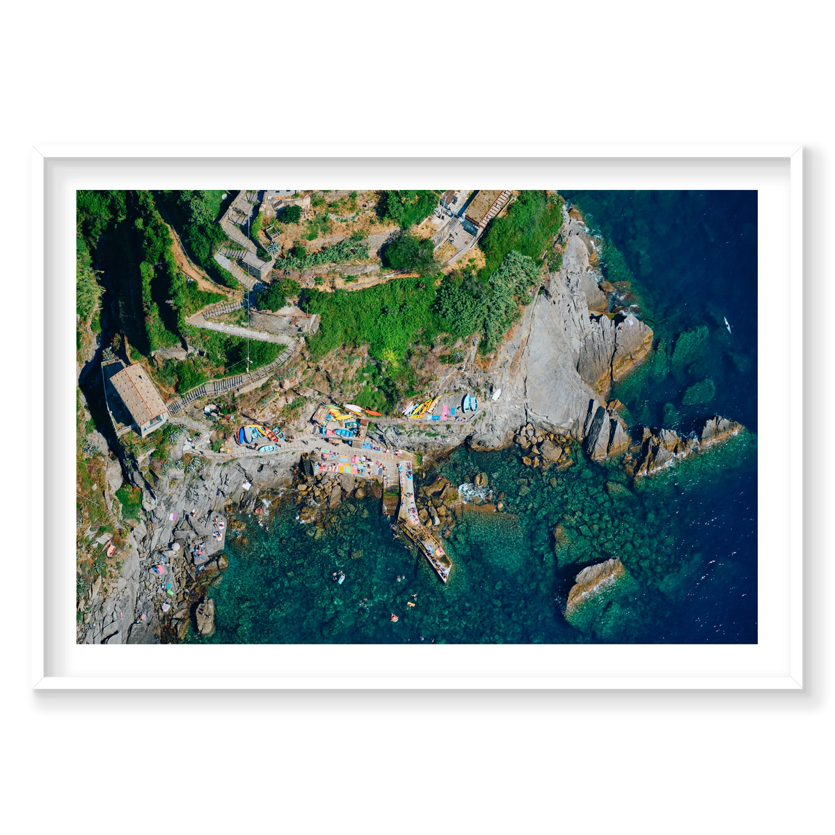 Overhead view of winding hillside steps, colourful boats, a rocky landing and swimmers in clear water at Corniglia, Cinque Terre, Italy, white frame.