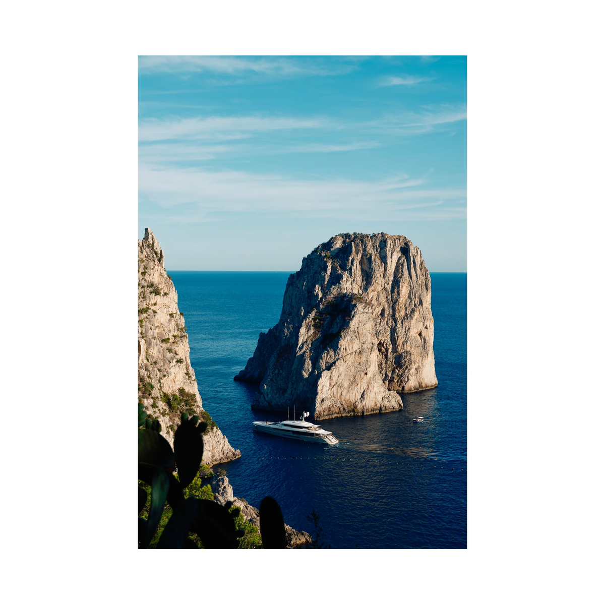 Endeavour 2 yacht beneath the cliffs of Capri, Italy, with blue sea, open sky and cactus in the foreground, print-only presentation.