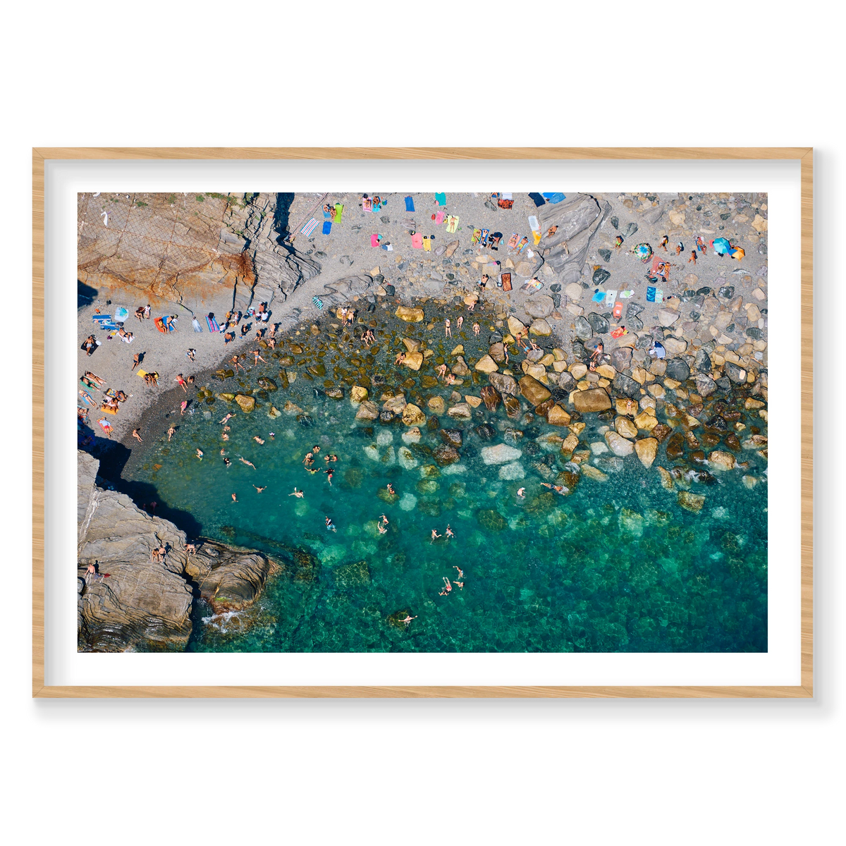 Overhead view of swimmers among boulders in clear green water, with bright towels on a rocky beach at Vernazza, Cinque Terre, Italy, natural timber frame.