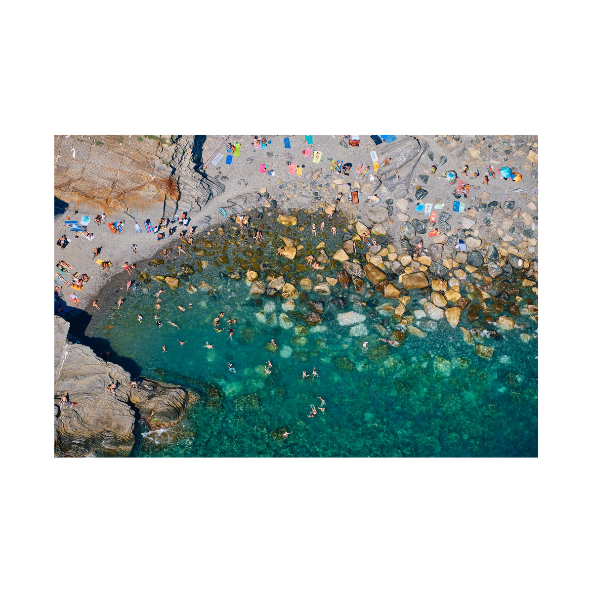 Overhead view of swimmers among boulders in clear green water, with bright towels on a rocky beach at Vernazza, Cinque Terre, Italy, print-only presentation.
