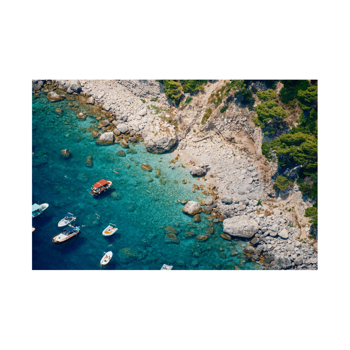 Swimmers and small boats in clear turquoise water beside the pale rocky shore of Capri, Italy, print-only presentation.
