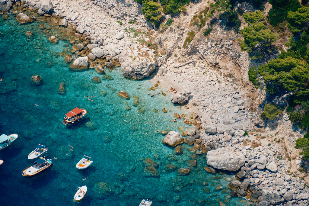 Swimmers and small boats in clear turquoise water beside the pale rocky shore of Capri, Italy, print-only presentation.