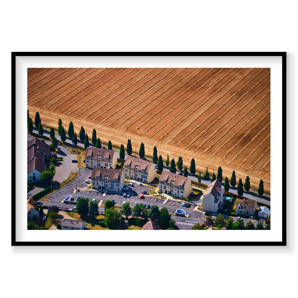 A row of houses and tall trees beside a harvested golden field in the Paris region, photographed from above; black frame