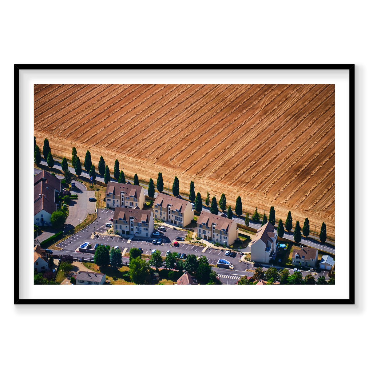 A row of houses and tall trees beside a harvested golden field in the Paris region, photographed from above; black frame