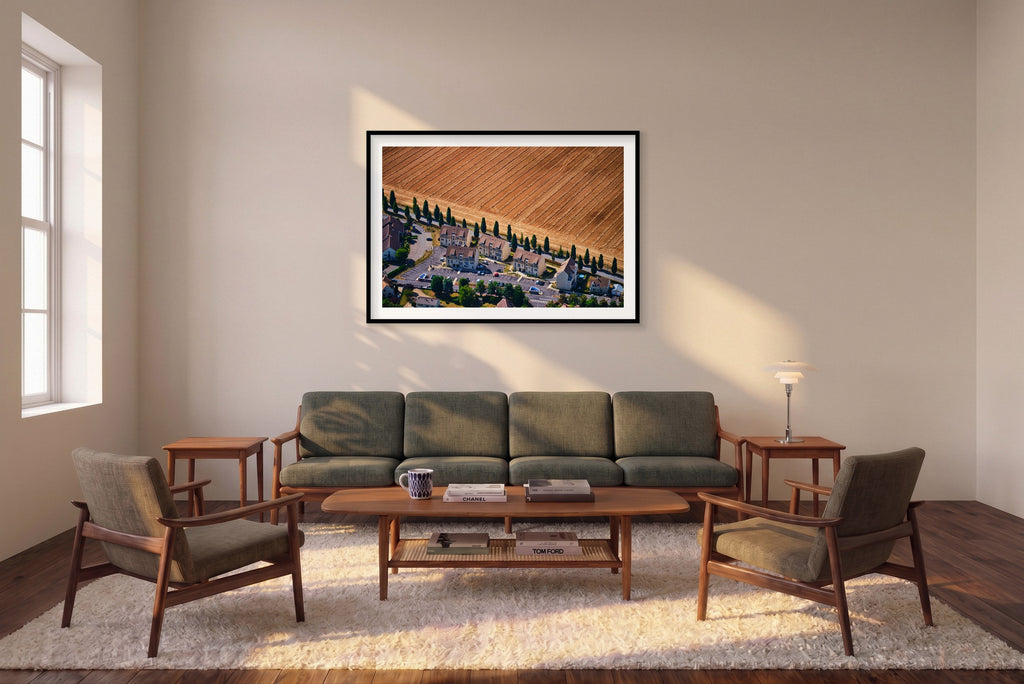 A row of houses and tall trees beside a harvested golden field in the Paris region, photographed from above; black framed print in a room setting