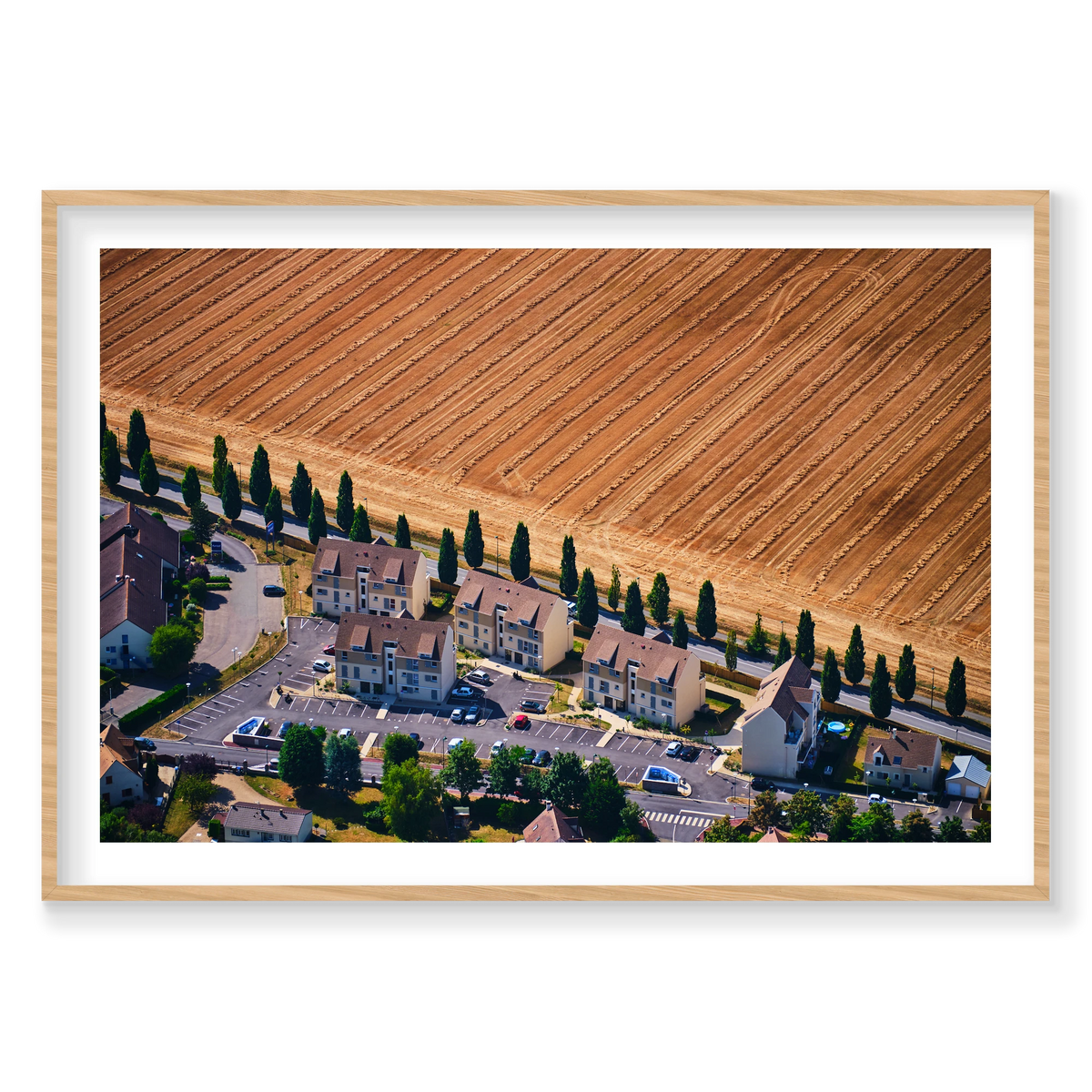 A row of houses and tall trees beside a harvested golden field in the Paris region, photographed from above; natural timber frame