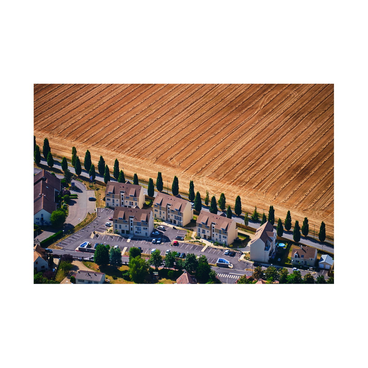 A row of houses and tall trees beside a harvested golden field in the Paris region, photographed from above; print only
