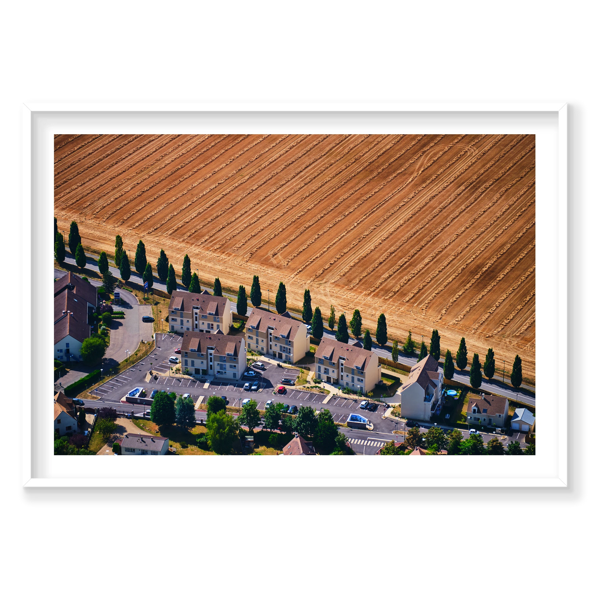 A row of houses and tall trees beside a harvested golden field in the Paris region, photographed from above; white frame
