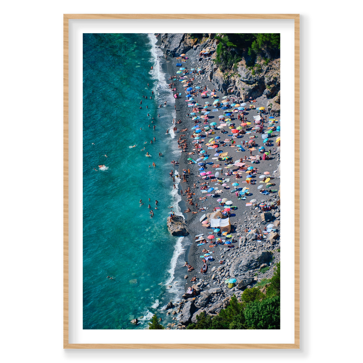 Colourful umbrellas and swimmers along the dark beach beneath rocky cliffs at Punta Corvo, Montemarcello, Italy, natural timber frame.