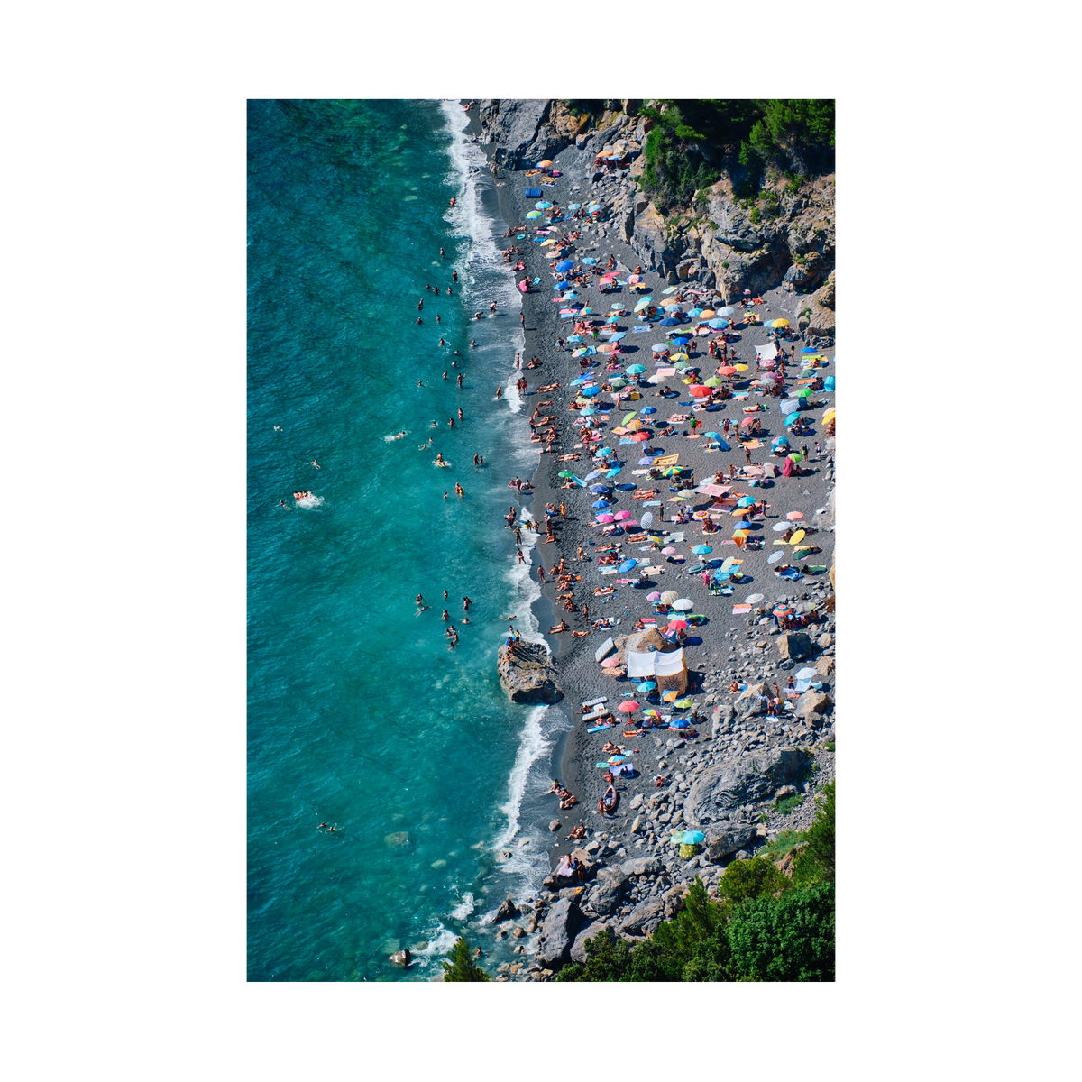 Colourful umbrellas and swimmers along the dark beach beneath rocky cliffs at Punta Corvo, Montemarcello, Italy, print-only presentation.