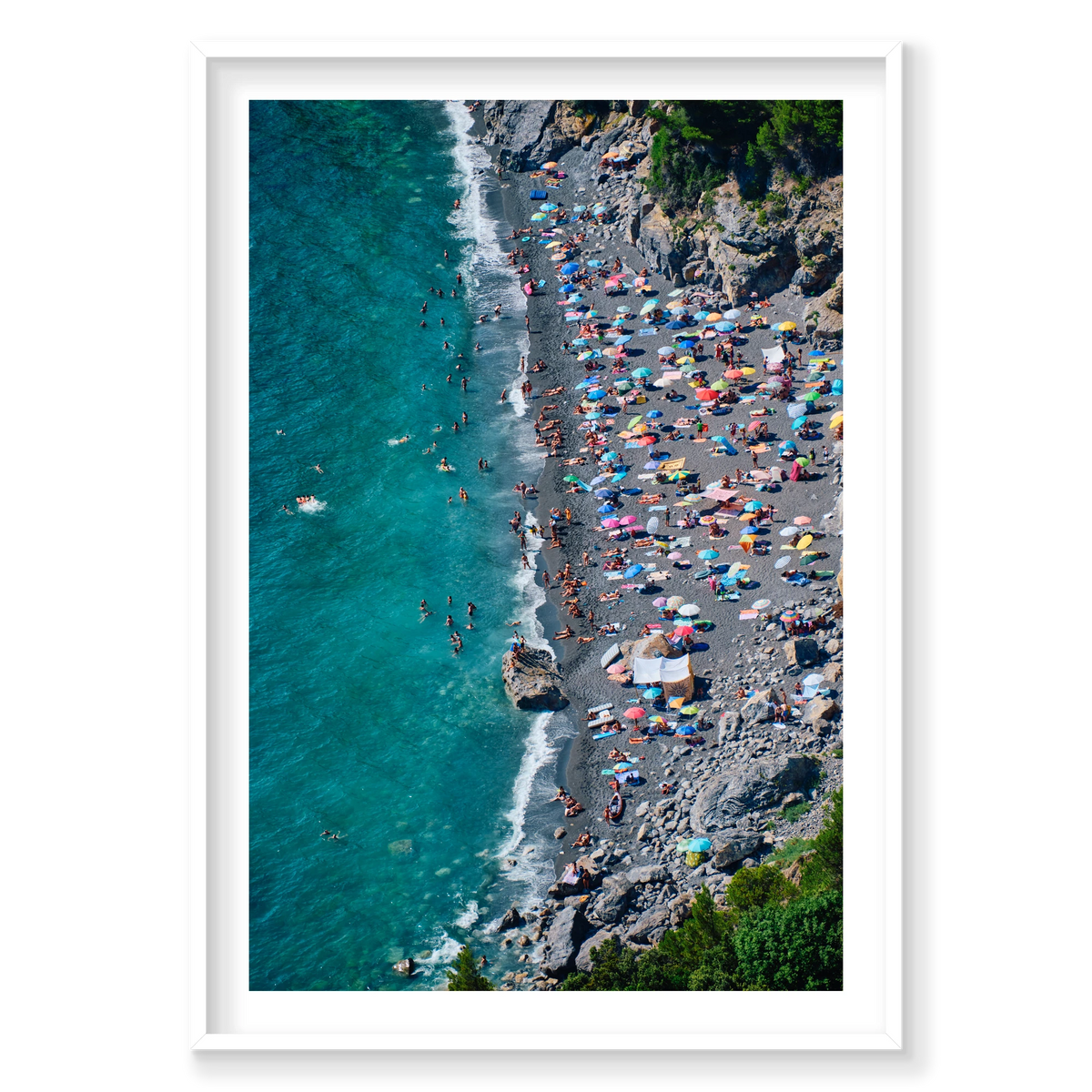 Colourful umbrellas and swimmers along the dark beach beneath rocky cliffs at Punta Corvo, Montemarcello, Italy, white frame.