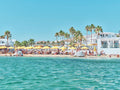 Mykonos beach view from turquoise water showing white buildings, palm trees, and yellow umbrellas, photography print by Re...