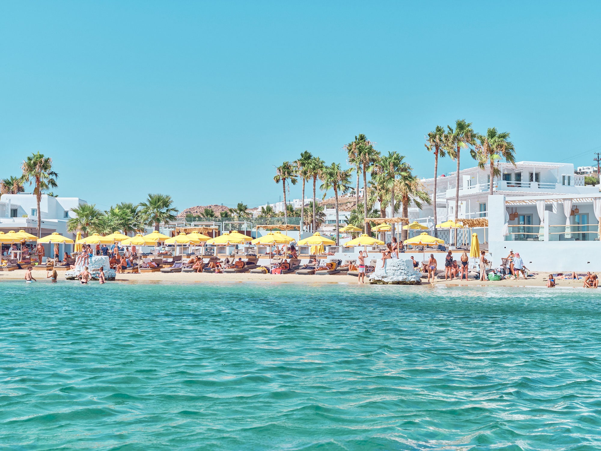 Mykonos beach view from turquoise water showing white buildings, palm trees, and yellow umbrellas, photography print by Re...