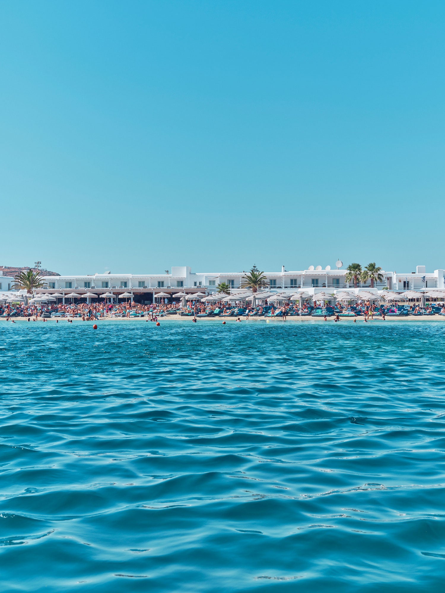 Ornos Beach Greece viewed from turquoise water with white buildings and beach umbrellas, photography print by Remy Gerega